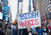 Person holding "Biden Harris" sign in crowded city street.