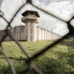 Alligator Alcatraz UNDER ATTACK — Activists Want SHUTDOWN…. View of prison watchtower through chain-link fence.