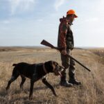 SHOCKING Proposal: Hunting Now a Crime? A man in hunting gear walking with a dog in a field during sunset
