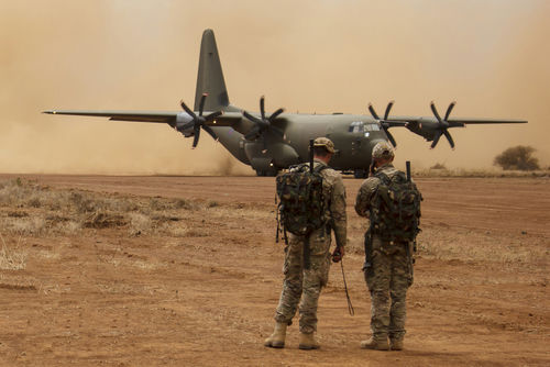 Two soldiers watch a military transport plane on a dusty airstrip