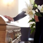 Person holding lilies beside a wooden coffin.