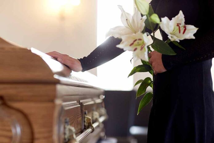 Person holding lilies beside a wooden coffin.