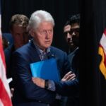 Bill Clinton standing between American flags with a folder.